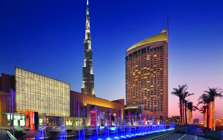 A modern hotel with a prominent tower, a large building with patterned facade, and a well-lit fountain area.