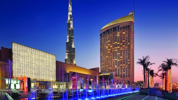 A modern hotel with a prominent tower, a large building with patterned facade, and a well-lit fountain area.