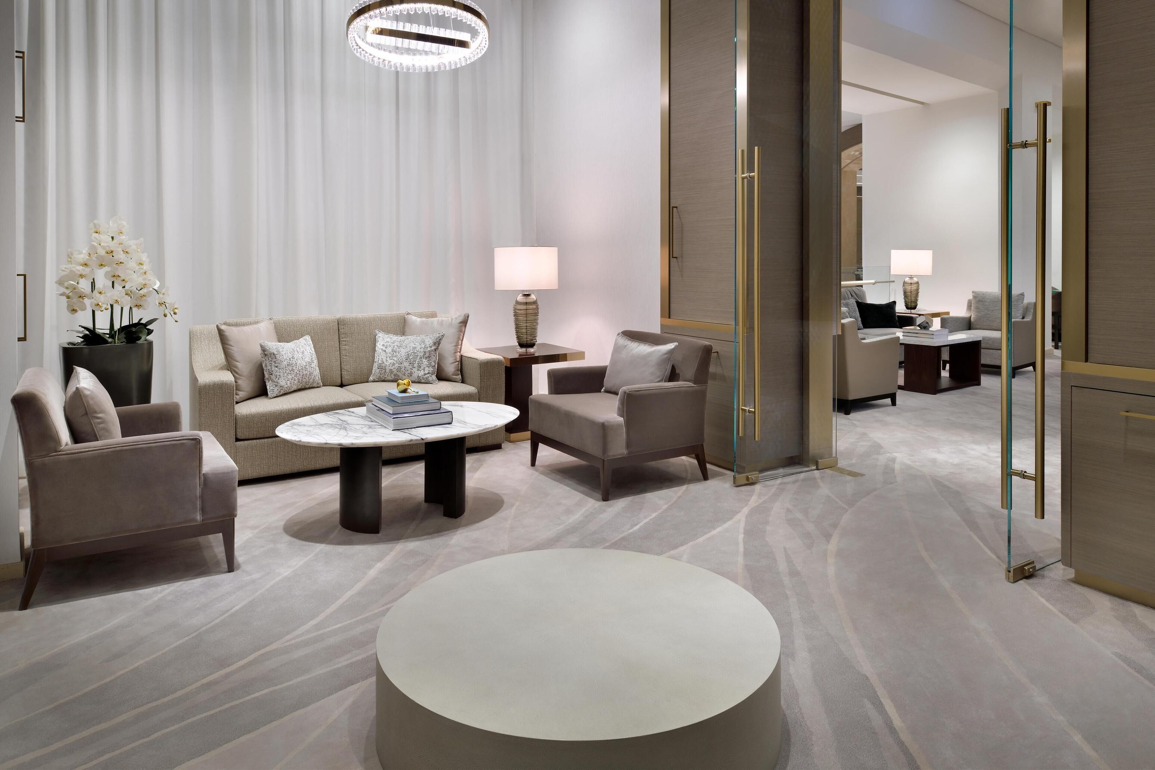 A modern hotel room with a beige sofa, two armchairs, a round coffee table, and a lamp.