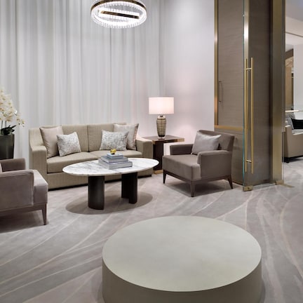 A modern hotel room with a beige sofa, two armchairs, a round coffee table, and a lamp.