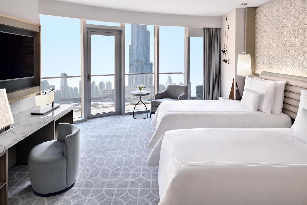 A hotel room with two beds, a desk, a chair, a small table, and a TV.