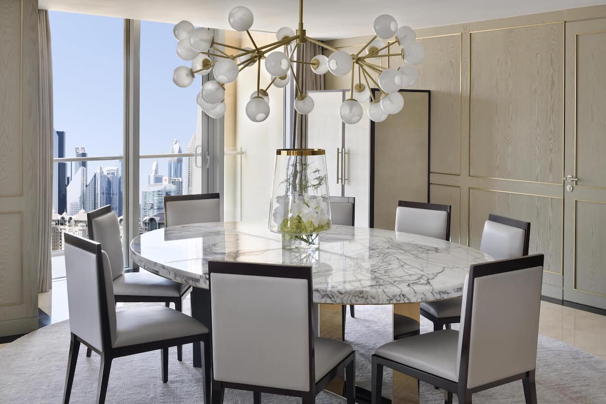A modern dining room with a marble table, six chairs, a chandelier, and a view of the city skyline.