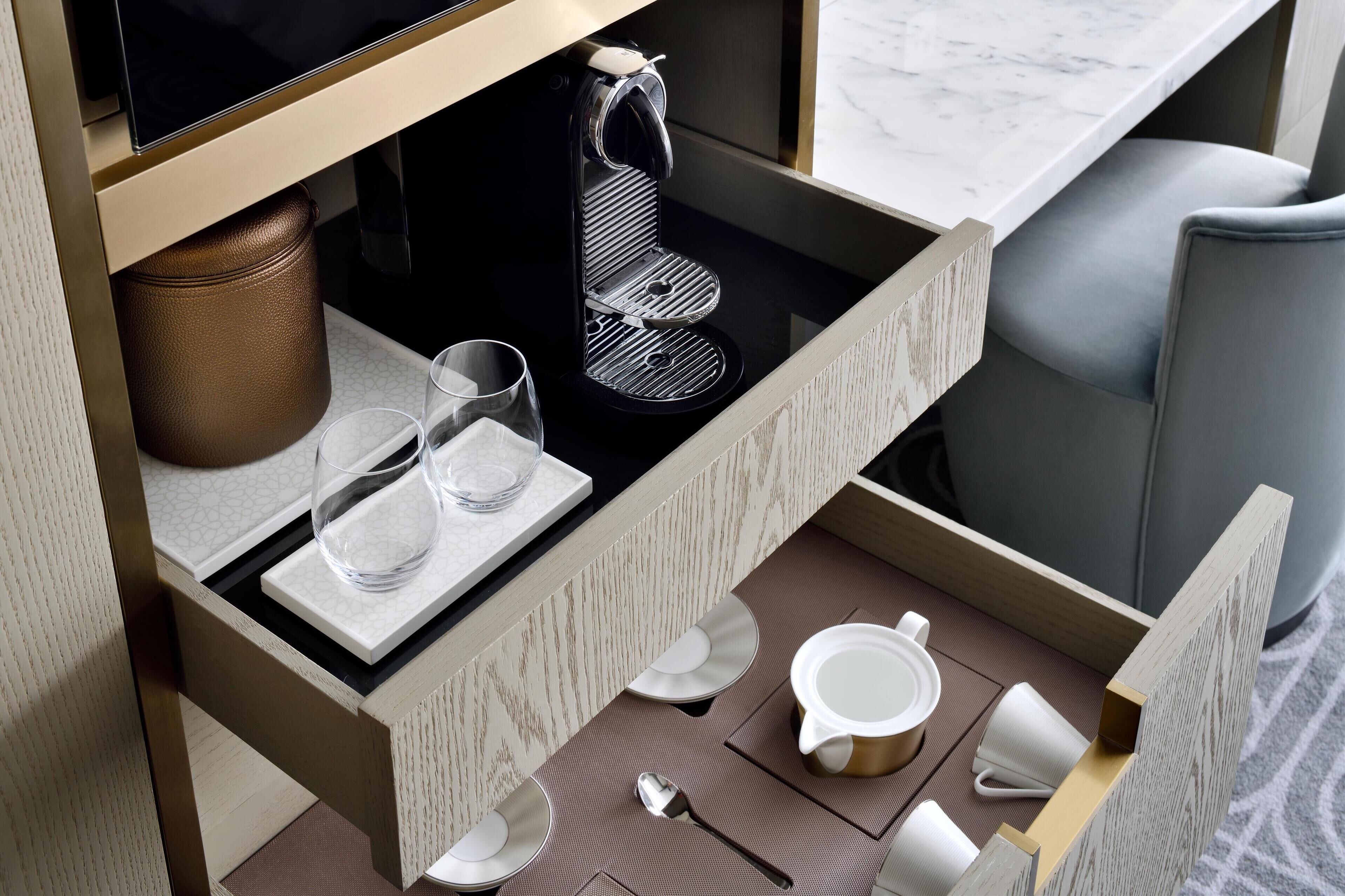 Habitación de hotel ordenada con cafetera, vasos, platos y una taza sobre la mesa.