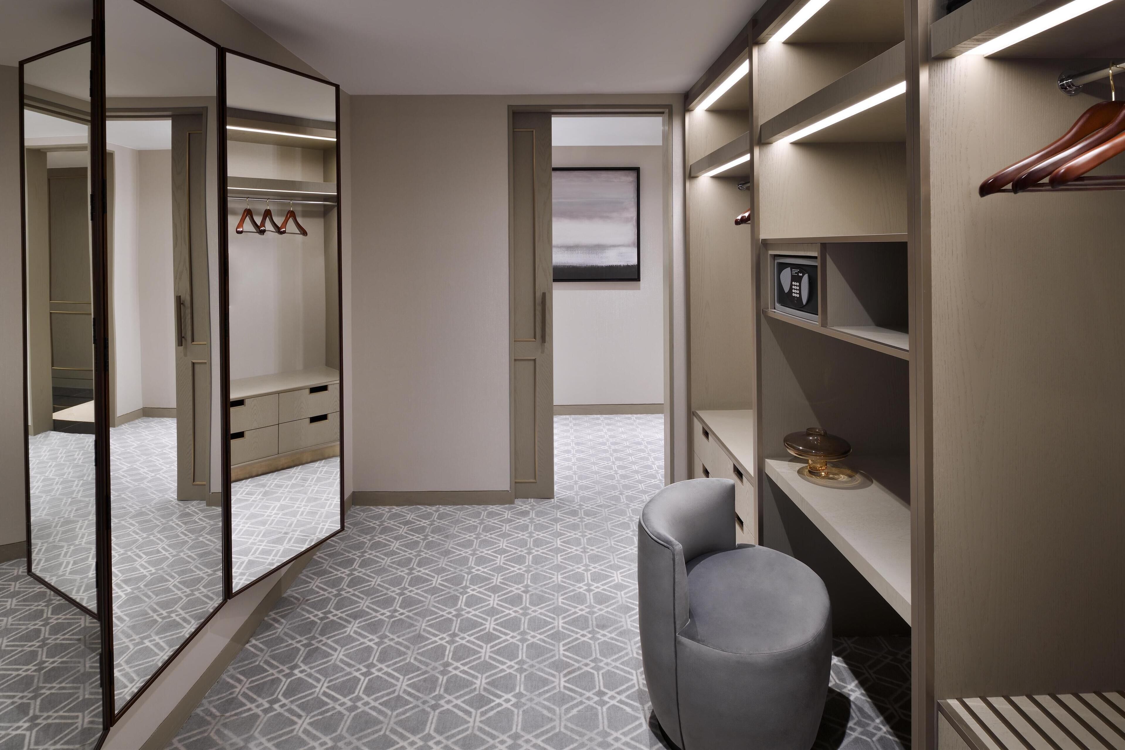 A walk-in closet with mirrored doors, a grey upholstered stool, and a window with a view.