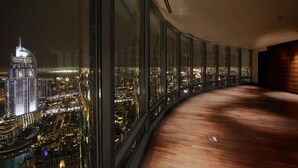 A high-rise hotel room with a curved wooden floor, large floor-to-ceiling windows offering a cityscape view, and a modern interior design.