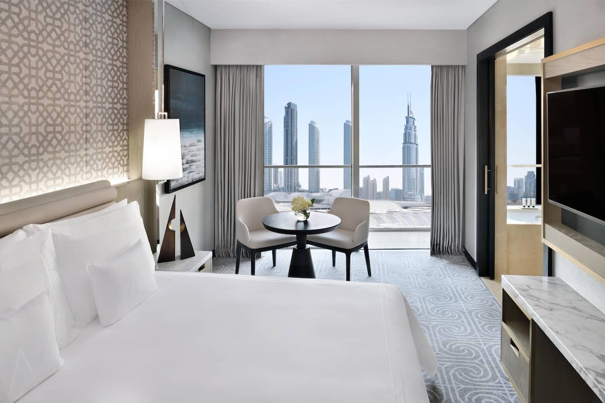 A hotel room with a large bed, a desk with a chair, a round table with a vase, and a city view through large windows.