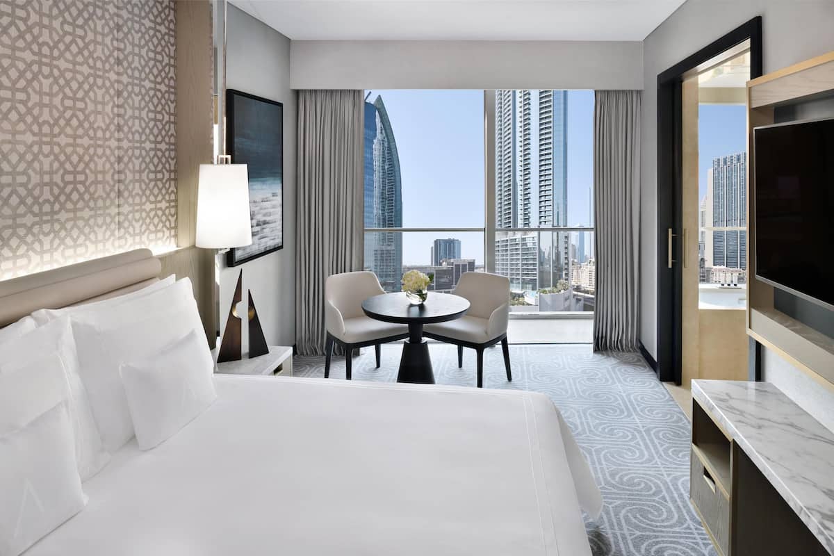 A hotel room with a large bed, a desk with a chair, a round table with a vase of flowers, and a large window offering a city view.