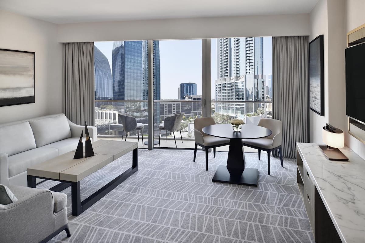 A modern hotel room with a large balcony, a sofa, a dining table, and a TV.