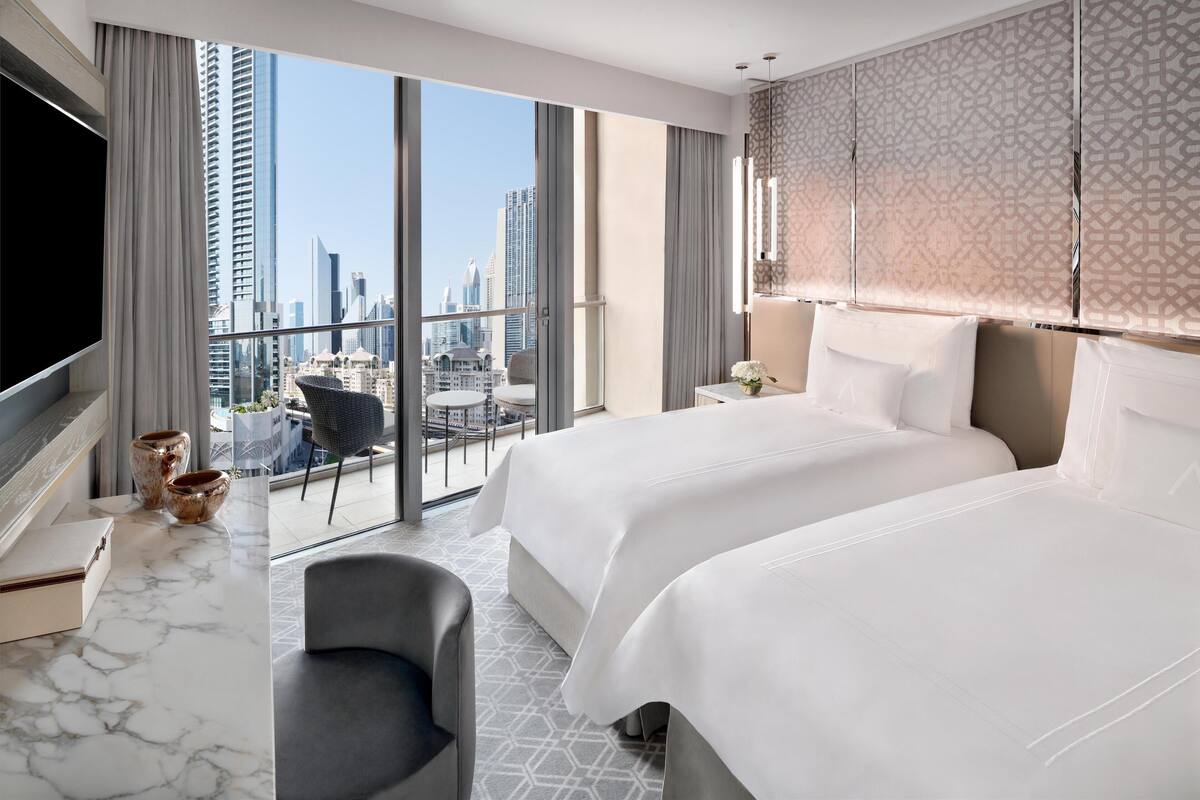 A hotel room with two beds, a large window with a city view, a marble desk, and a modern chair.