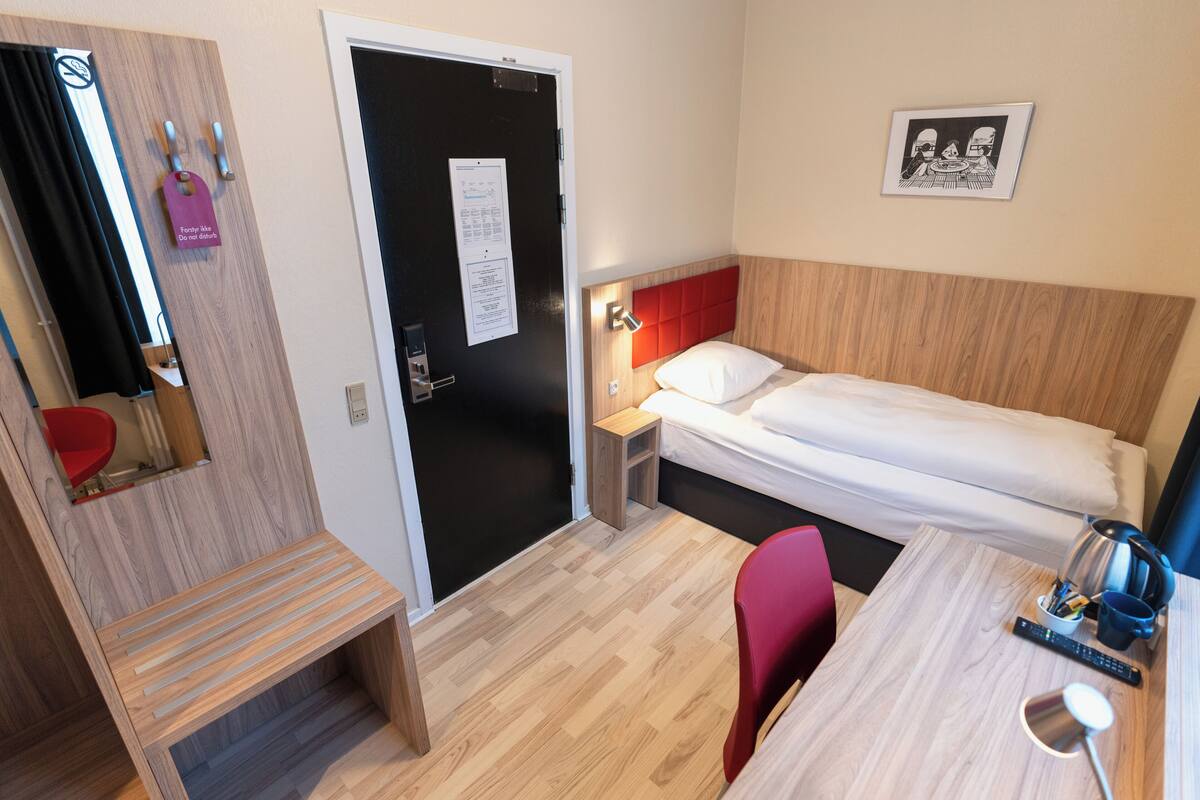 A hotel room with a bed, bedside table, chair, and a small desk with a lamp.