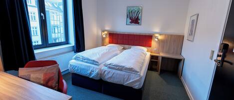 A hotel room with a bed, bedside tables, a desk, a chair, a window with curtains, and artwork on the walls.