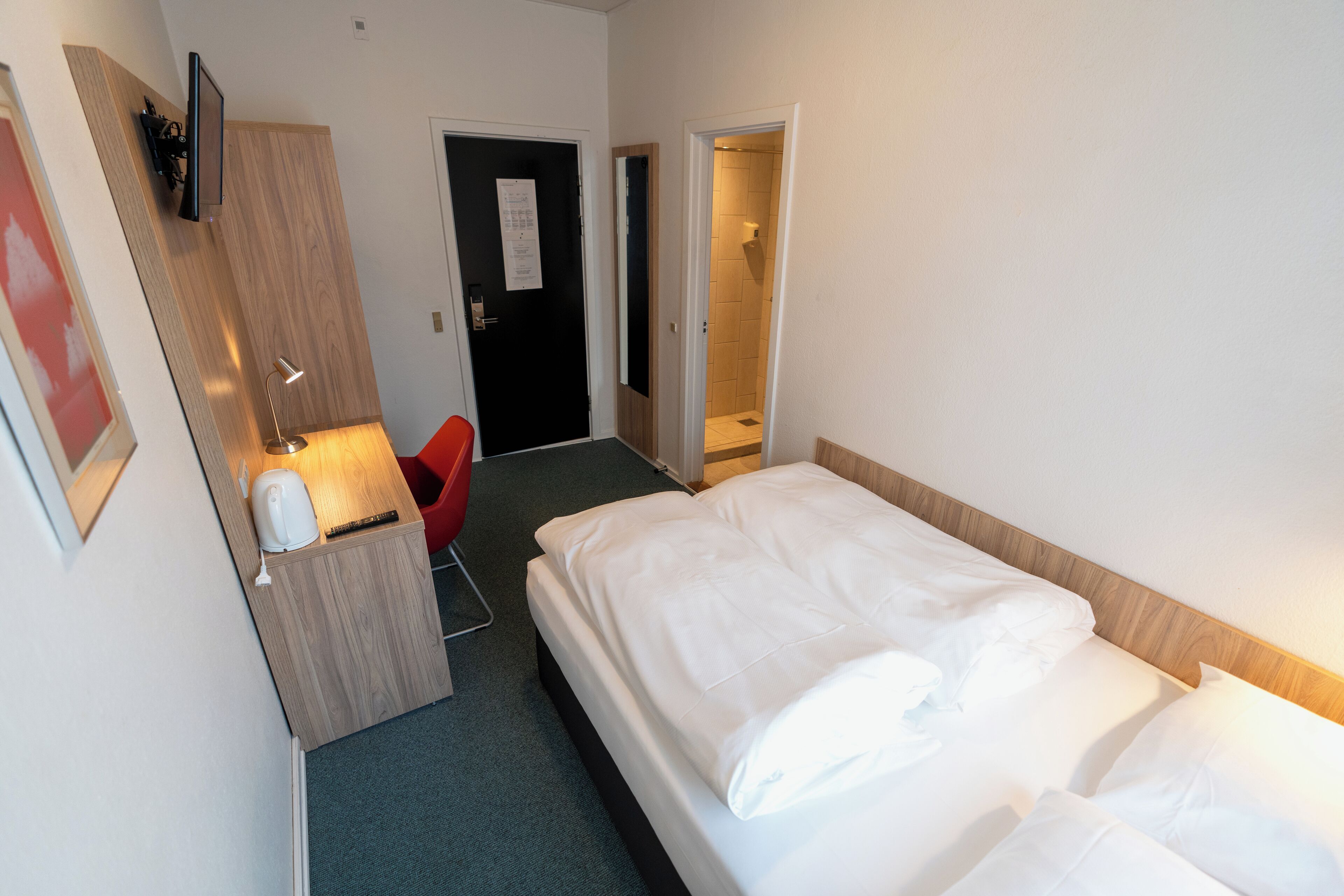 Habitación de hotel con cama, escritorio con lámpara, una silla roja y zona de ducha.