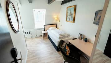 Habitación de hotel con cama, escritorio, silla, radiador, ventana y un cuadro en la pared.
