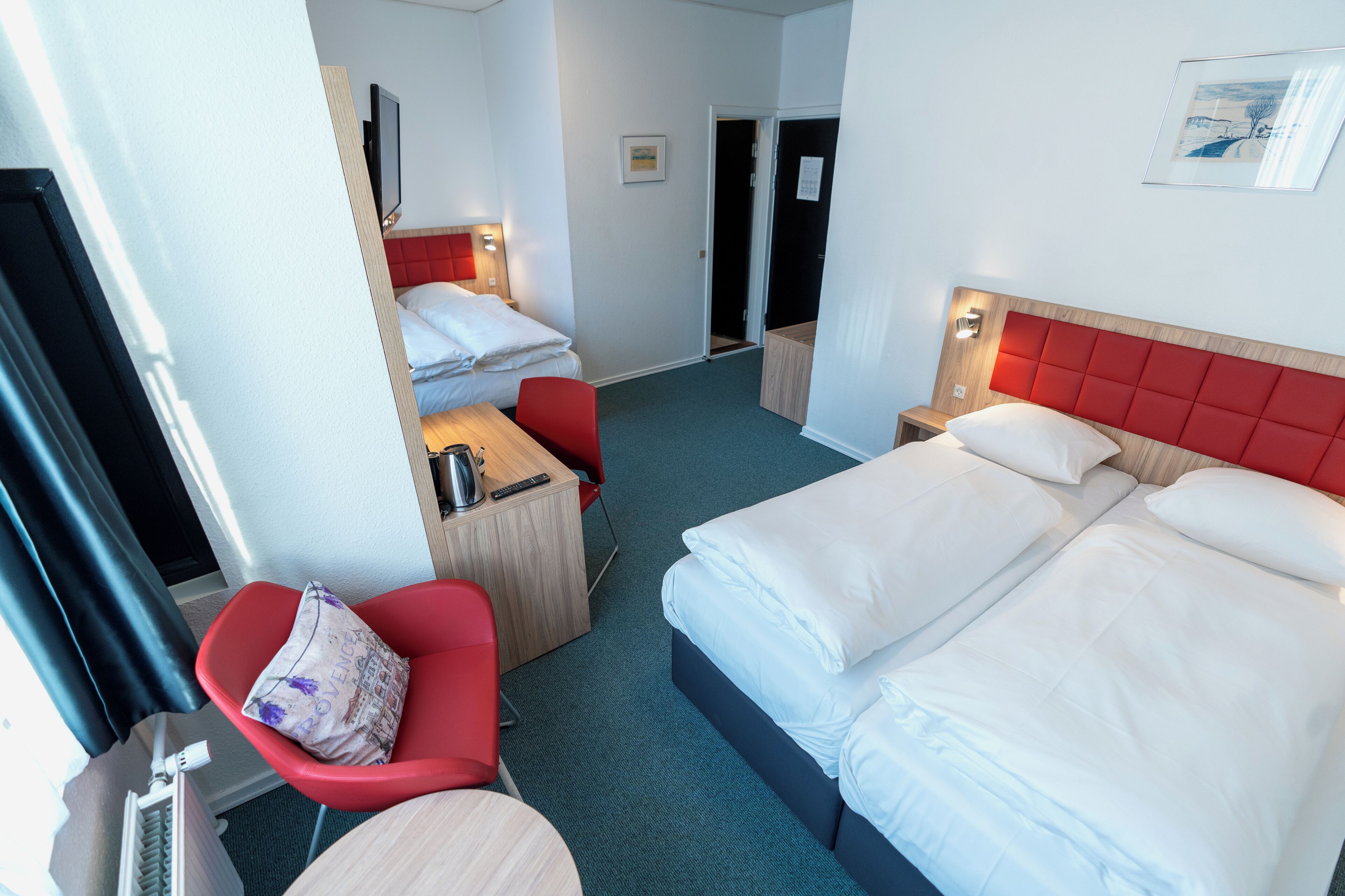A hotel room with a large bed, a desk with a chair, a television, and a red upholstered headboard.