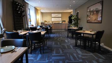 A hotel dining area with multiple tables and chairs, a kitchen in the background, and a large window providing natural light.