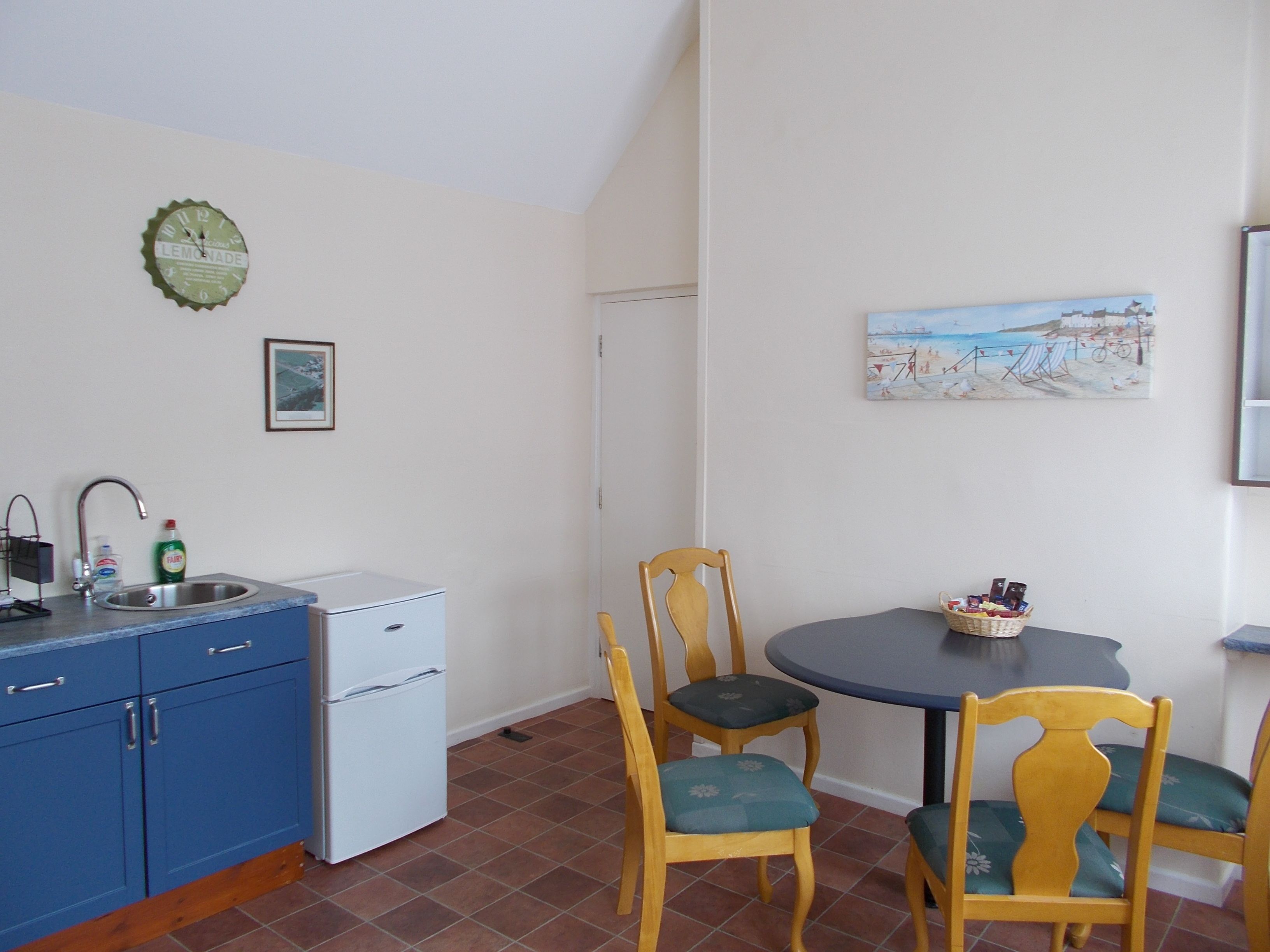 A compact kitchen with a sink, a small white refrigerator, and blue cabinets. There is a dining table with two chairs and a painting on the wall.