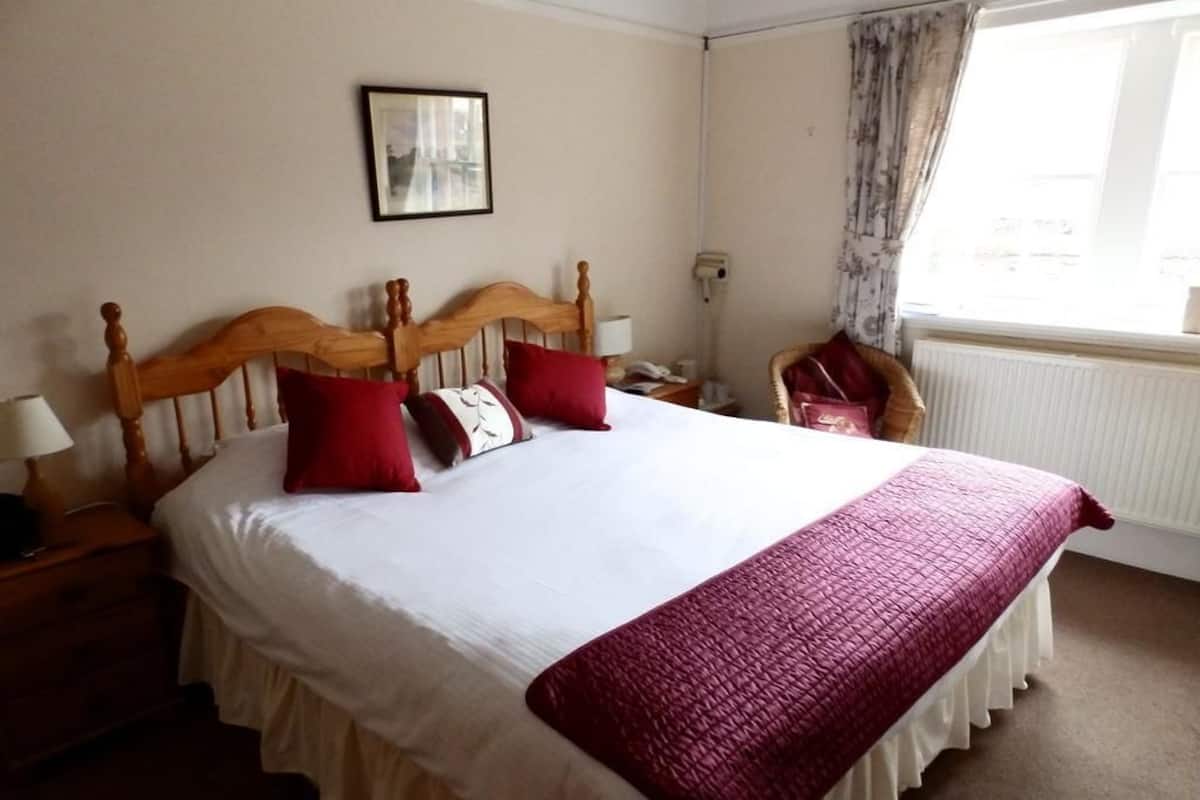 A neatly made bed with a wooden headboard, white bedding, and a red throw blanket. There is a nightstand with a lamp and a framed picture on the wall. A chair is visible in the corner, and a window with curtains is on the right.