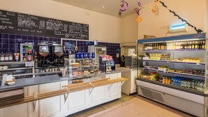 A cafe with a menu board, coffee machines, a display case with pastries, and shelves stocked with beverages. - The Coachman's Quarters at Muncaster Castle (Ravenglass)