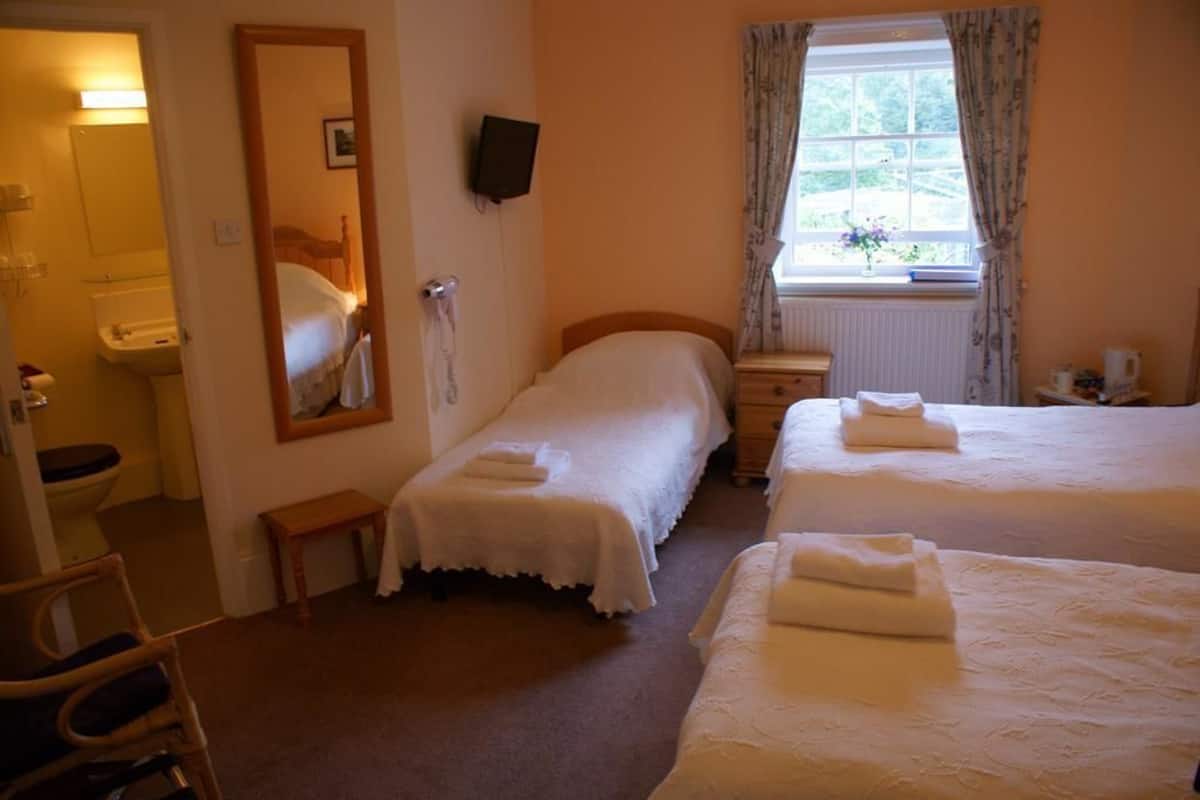 A hotel room with two beds, a bedside table, a chair, a mirror, a window with curtains, and a bathroom visible through an open door.