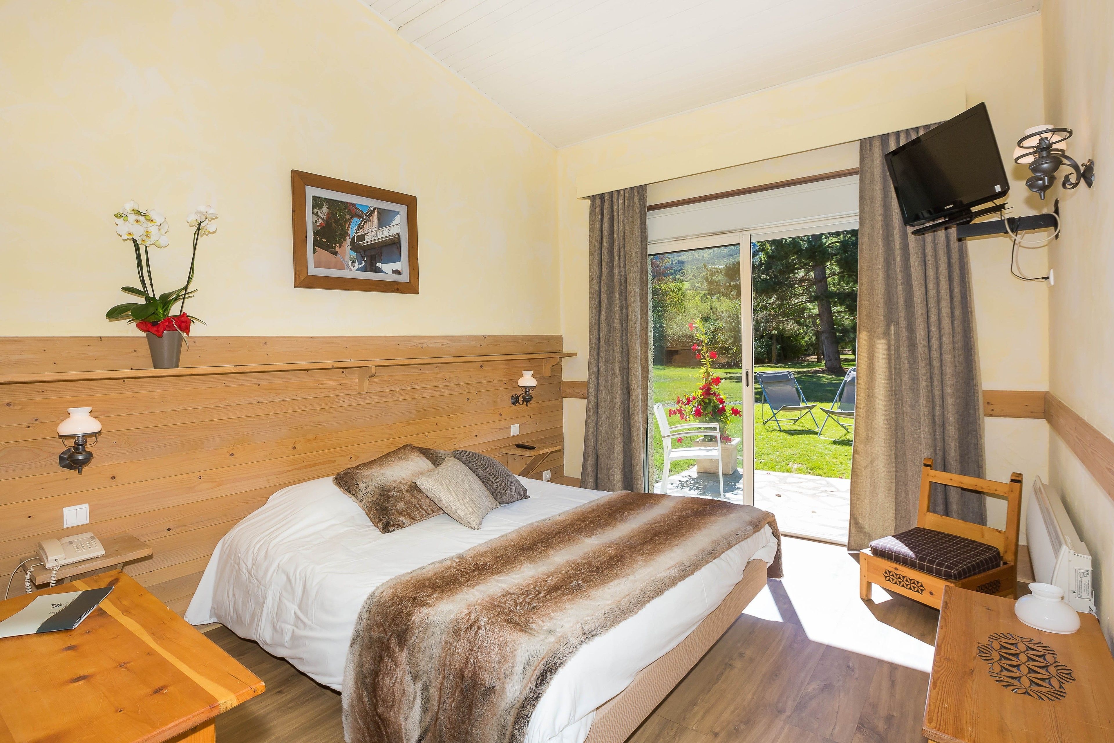 Une chambre d’hôtel avec une tête de lit en bois, un lit avec une literie blanche, une table de chevet en bois avec une lampe, une petite table avec une chaise, un téléviseur fixé au mur et une fenêtre donnant sur un jardin.