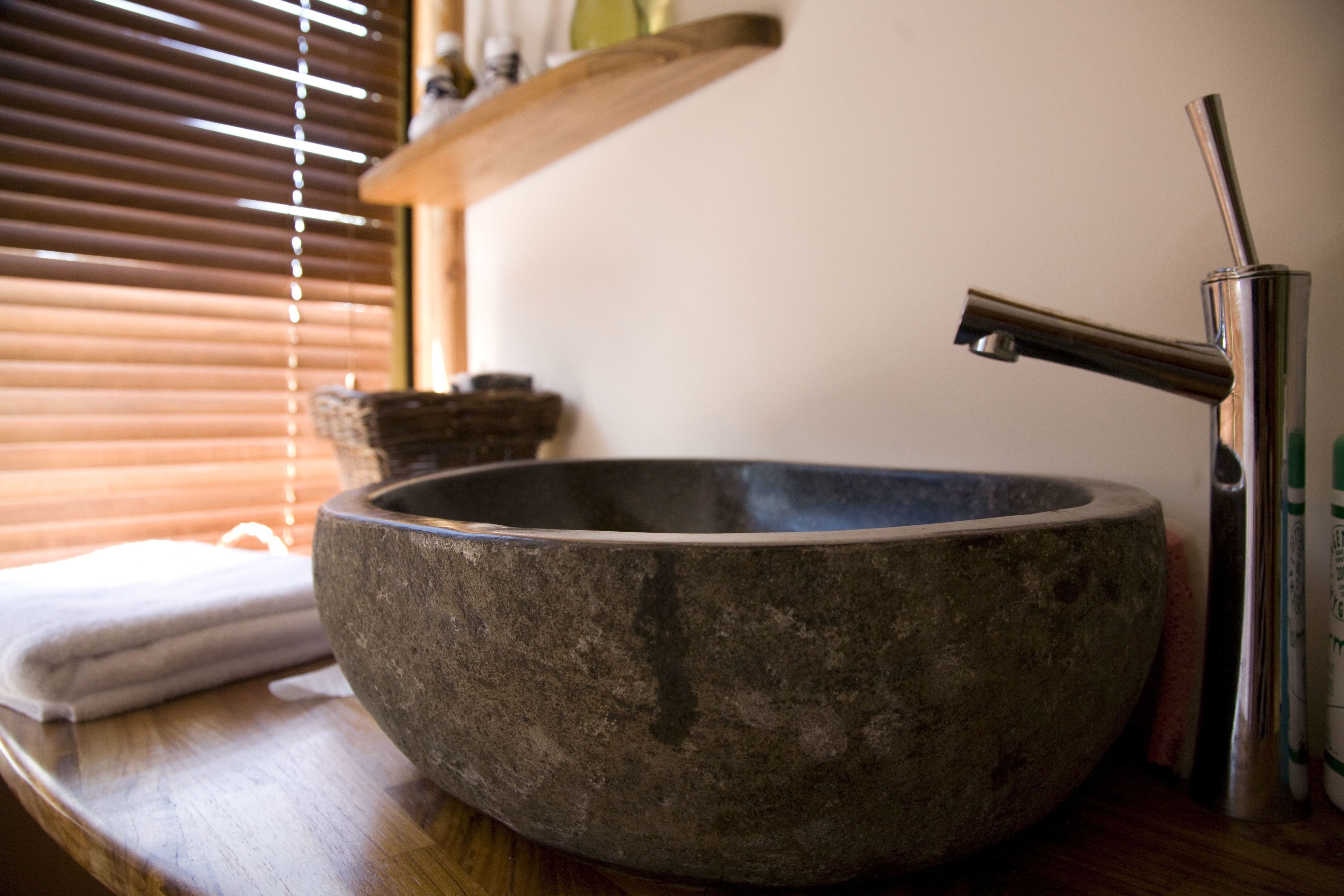 A stone basin with a modern faucet, a wooden shelf above, and a window with blinds in the background.