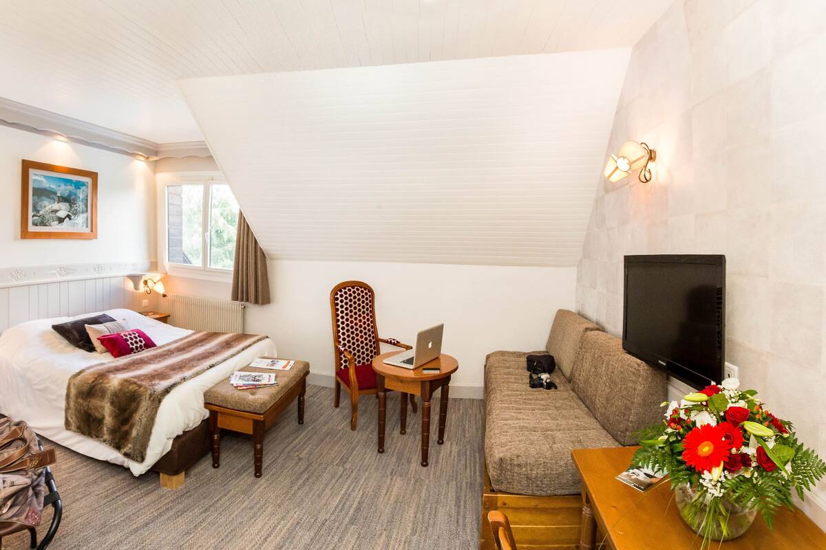 A cozy room with a bed, a desk with a laptop, a sofa, a TV, and a floral arrangement.