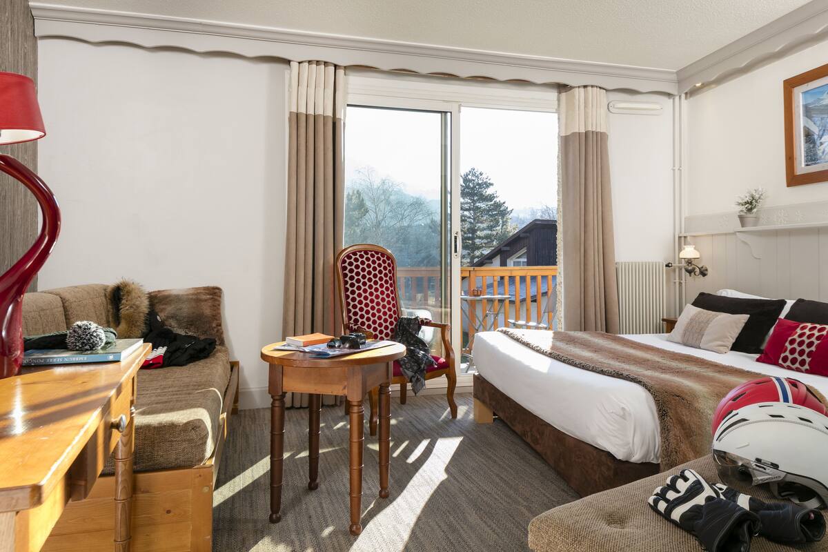 A hotel room with a bed, a sofa, a dining table, and a chair. There is a window with curtains, a radiator, and a plant on the windowsill.