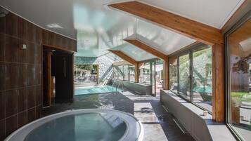 A modern indoor pool area with a hot tub, large windows, and a stone wall in the background.
