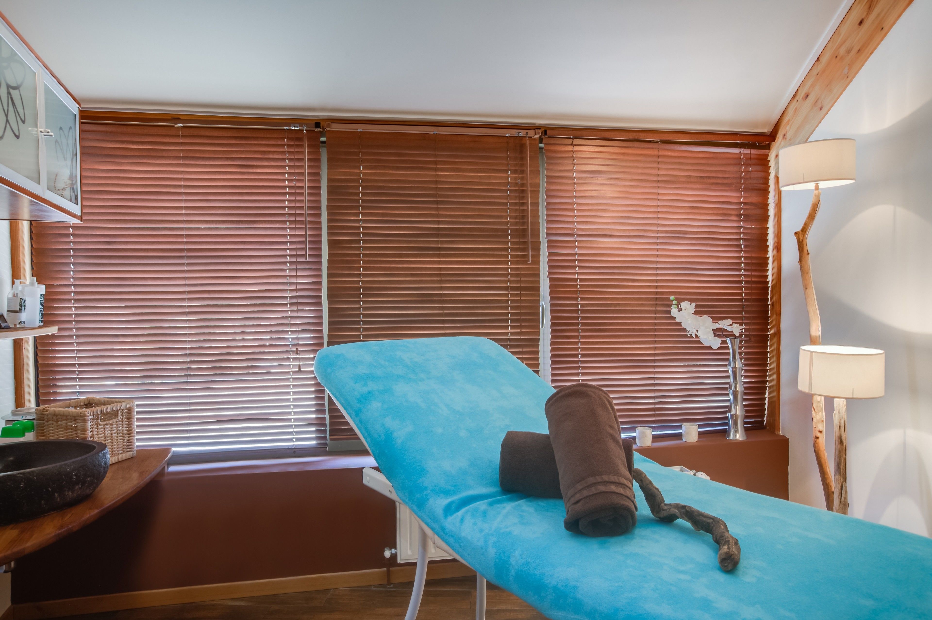 Una habitación con una camilla de masaje, persianas de madera, una lámpara y una mesita con una cesta y un cuenco.
