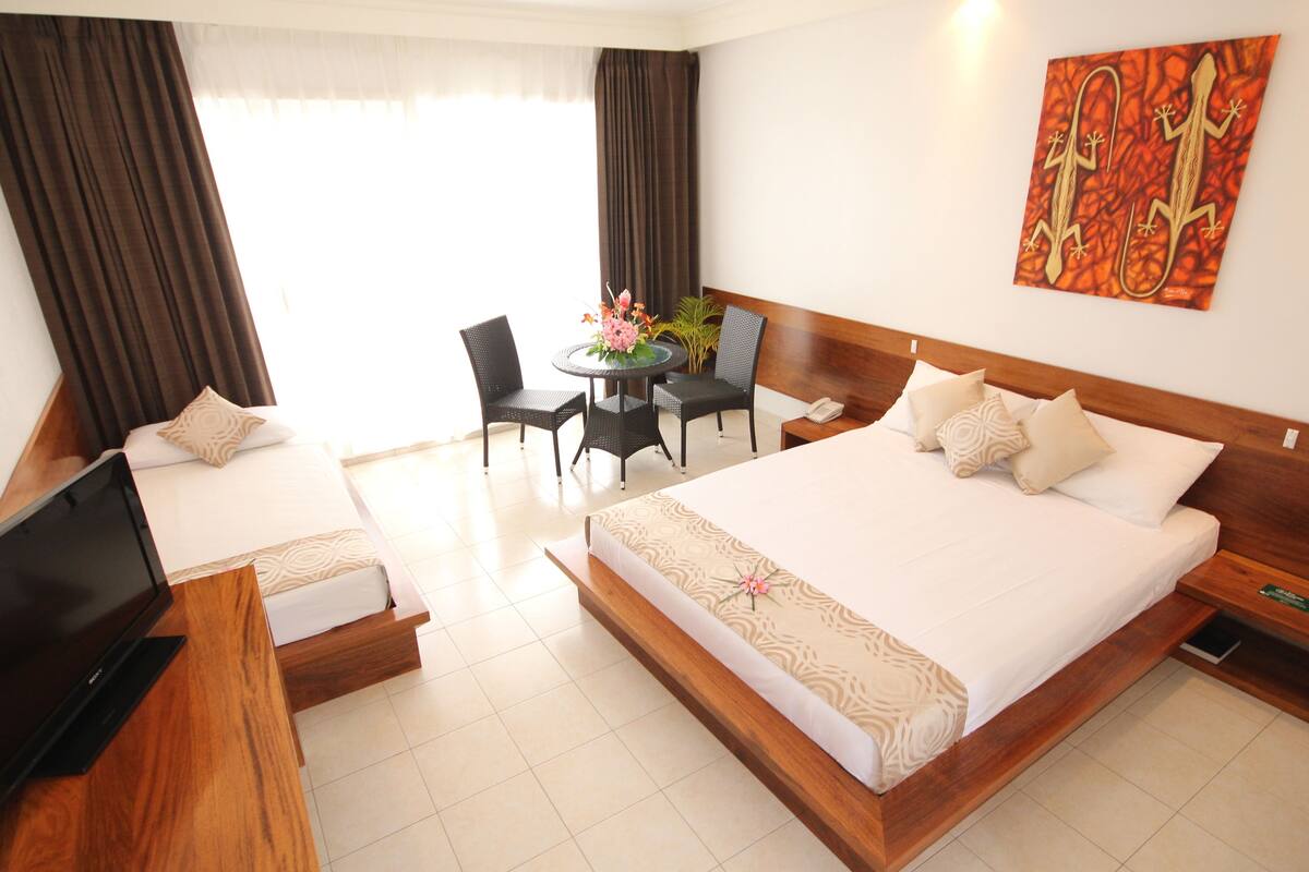 A hotel room with a large bed, a TV, a desk with a chair, a small table with flowers, and a painting on the wall.