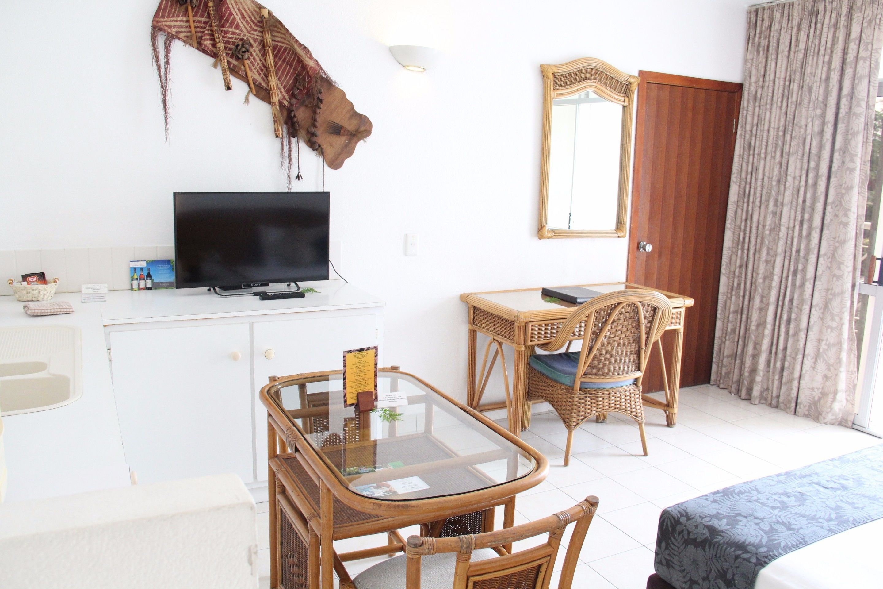 A hotel room with a glass-top dining table, a television on a white cabinet, a wicker chair, a bed with a patterned bedspread, a wooden door, and a large mirror.