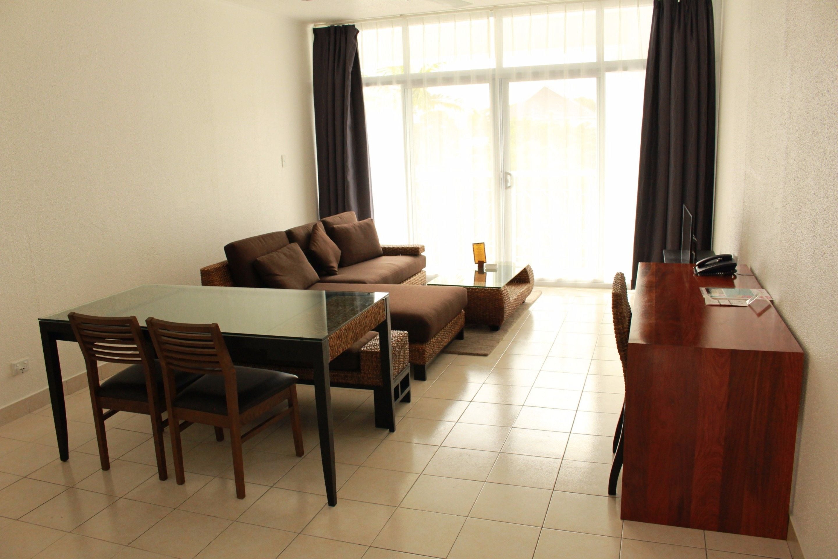 A living room with a glass-top dining table, brown sofa, and wooden chairs.