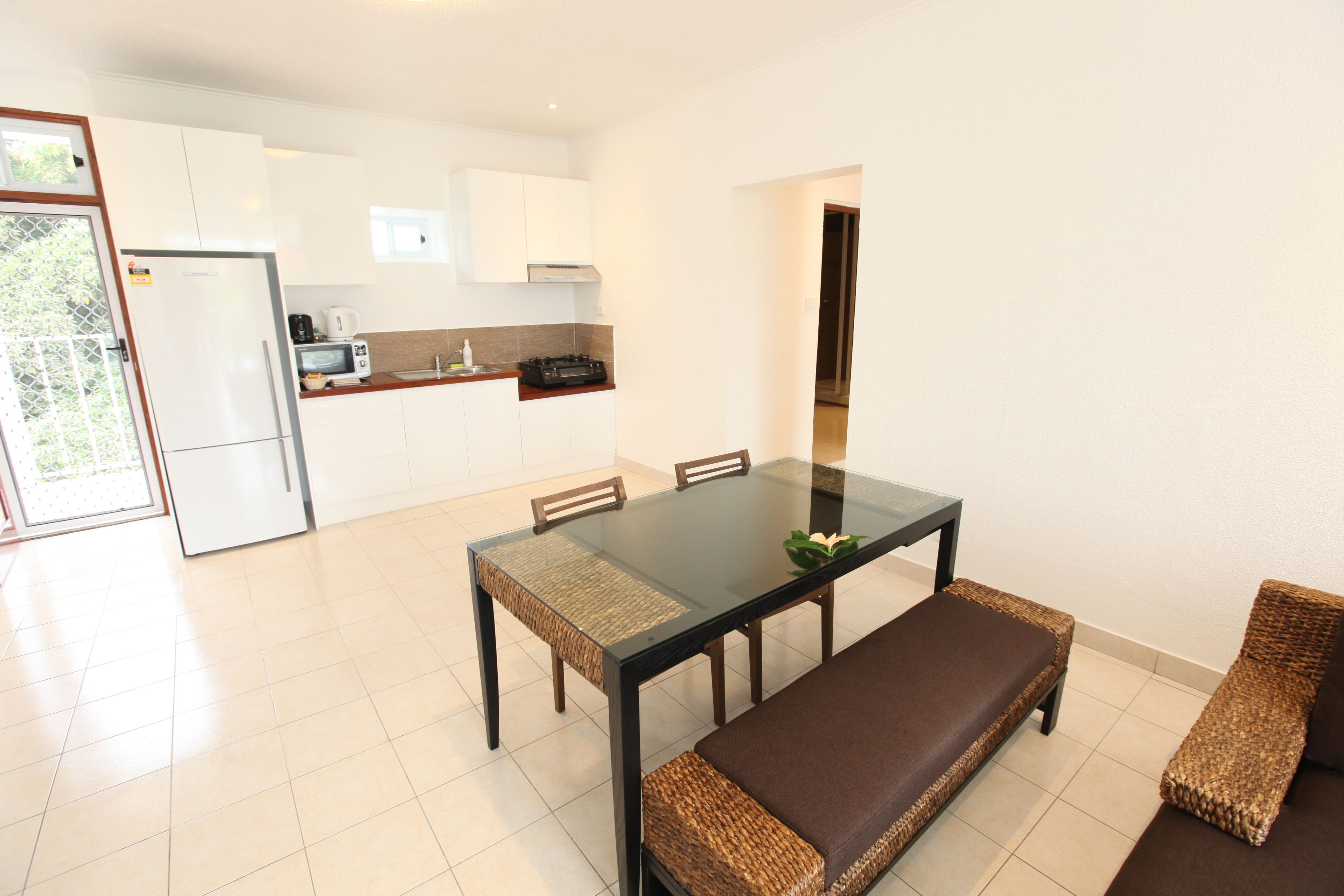 A modern kitchen with white cabinets, a refrigerator, microwave, and coffee maker. There is a glass dining table with four chairs and a bench with woven seats.