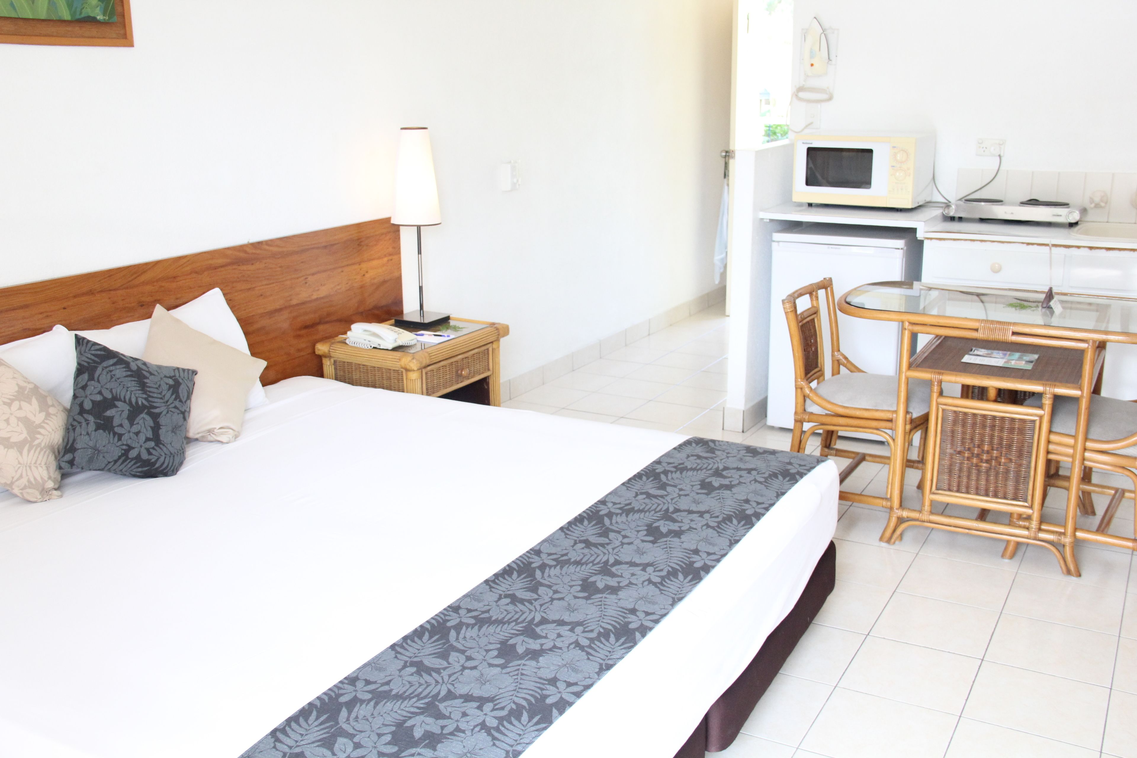 A hotel room with a bed, bedside lamp, wicker nightstand, microwave, and a small dining table with chairs.