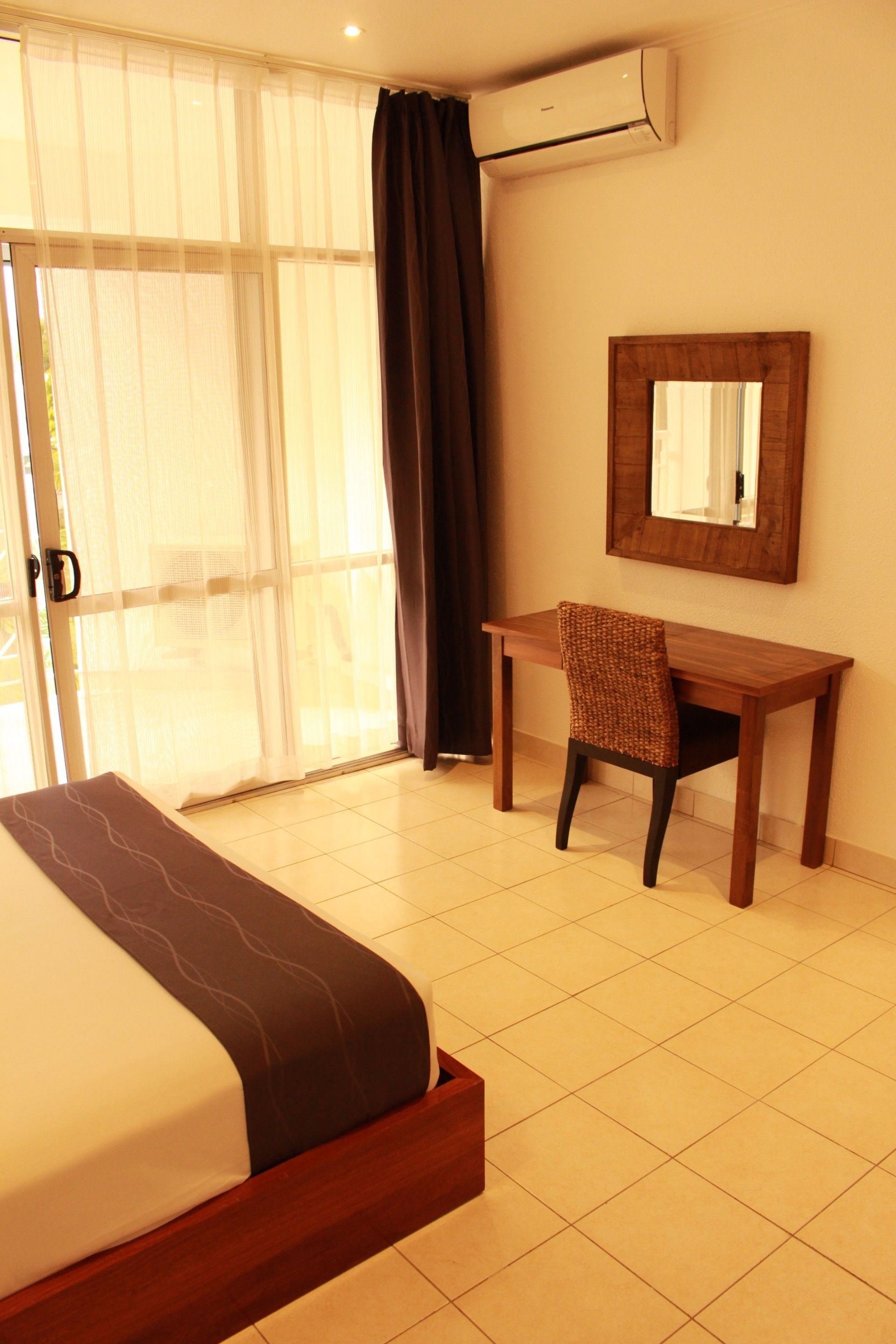 A hotel room with a bed, a desk, a chair, a mirror, and an air conditioning unit.
