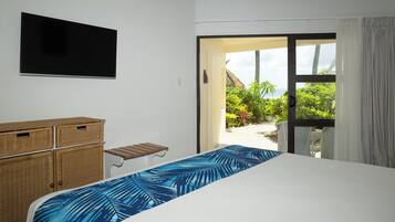 Une chambre d’hôtel avec un lit, un téléviseur fixé au mur, des meubles en osier et une vue sur un jardin tropical et une plage.
