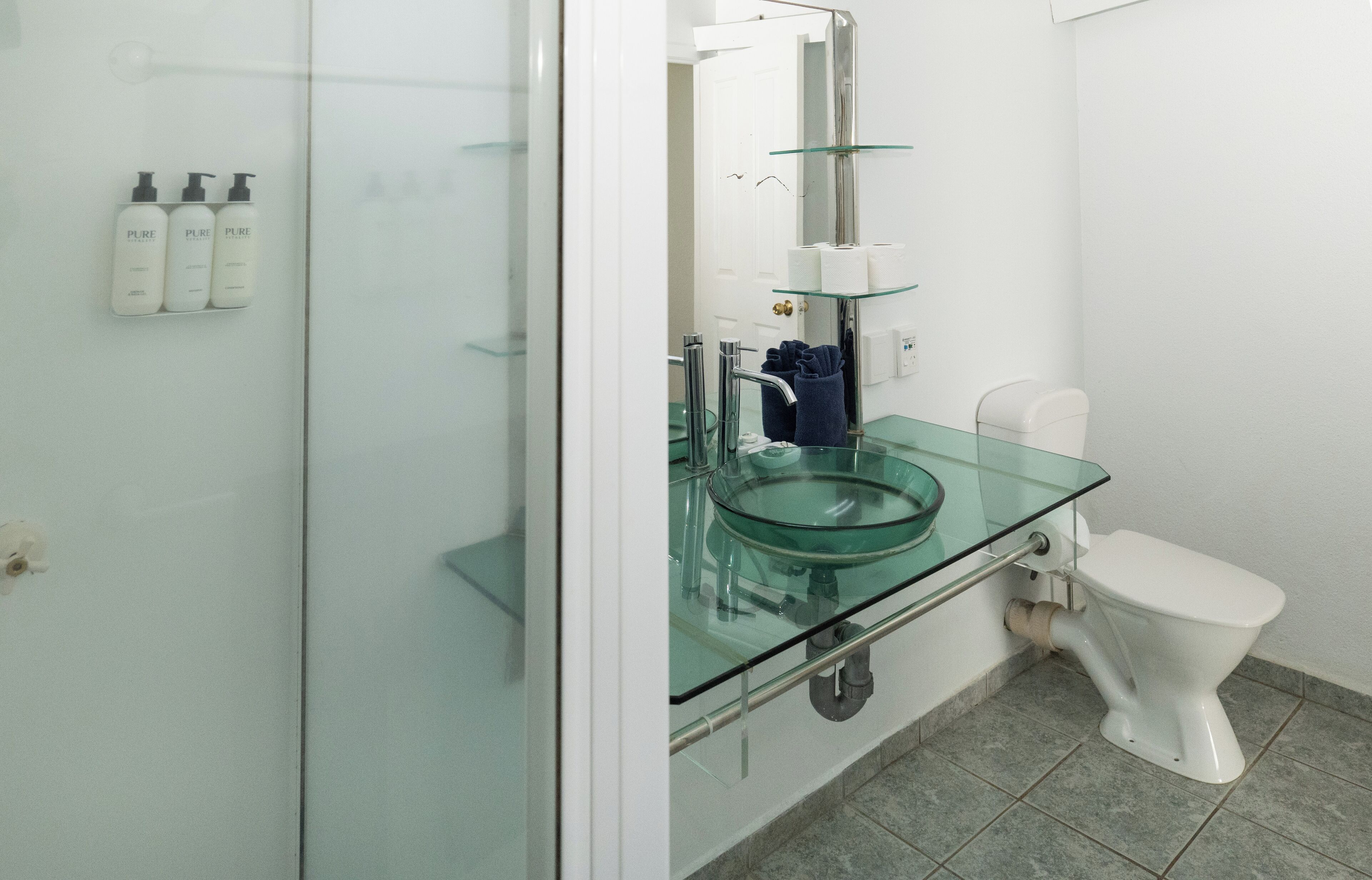 A modern bathroom with a glass sink, white toilet, and three bottles of Pure shampoo on the wall.