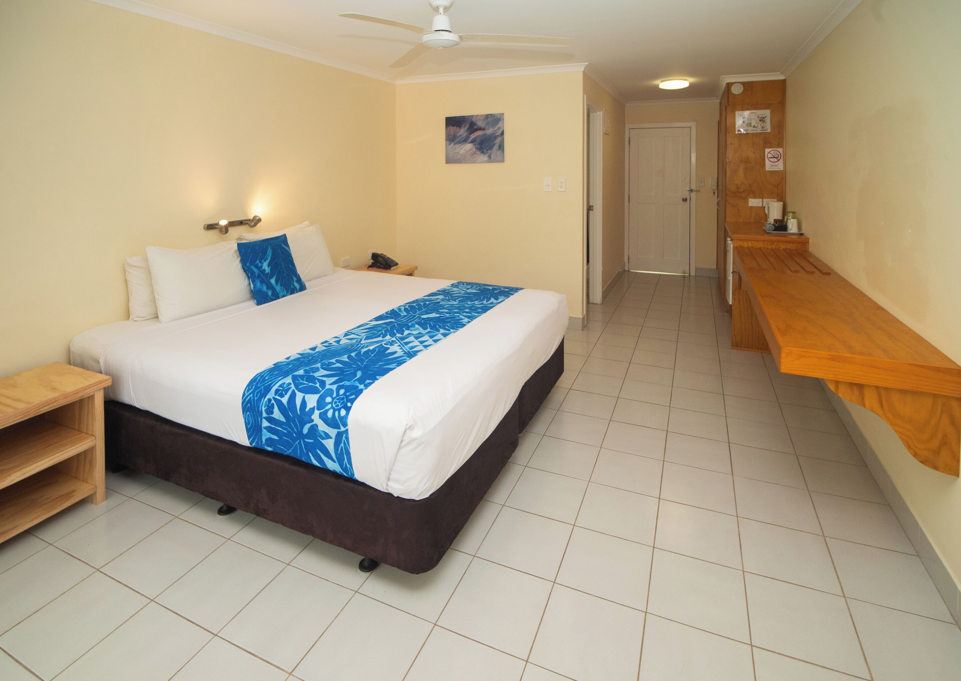 A hotel room with a bed, a wooden nightstand, a desk, a ceiling fan, and a door leading to another room.