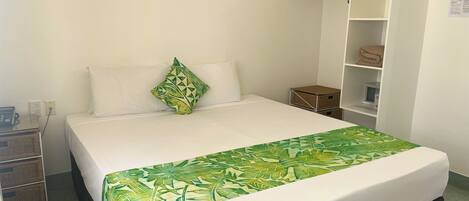 A neatly made bed with a green leaf-patterned bedspread, a white pillow, and a green geometric pillow. There is a white wardrobe with a shelf on top, a small white cabinet, and a wall-mounted shelf with folded towels.