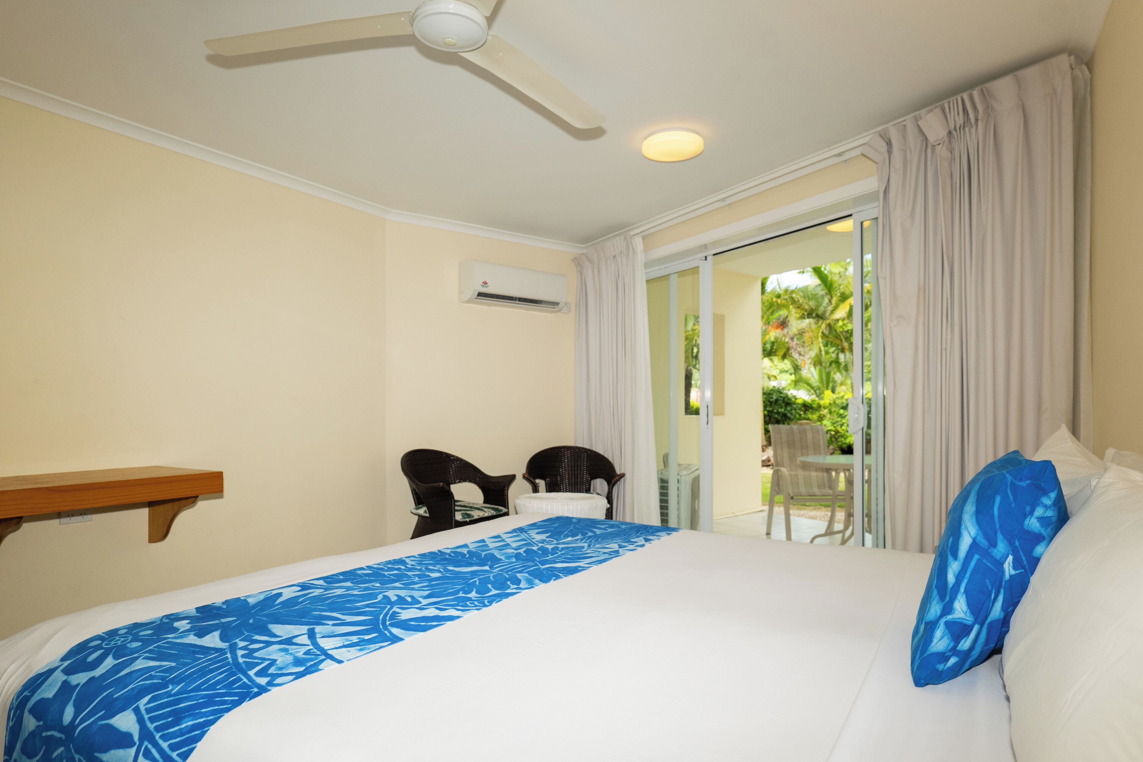 A hotel room with a bed, a ceiling fan, an air conditioning unit, a window with curtains, and a view of the outdoors.