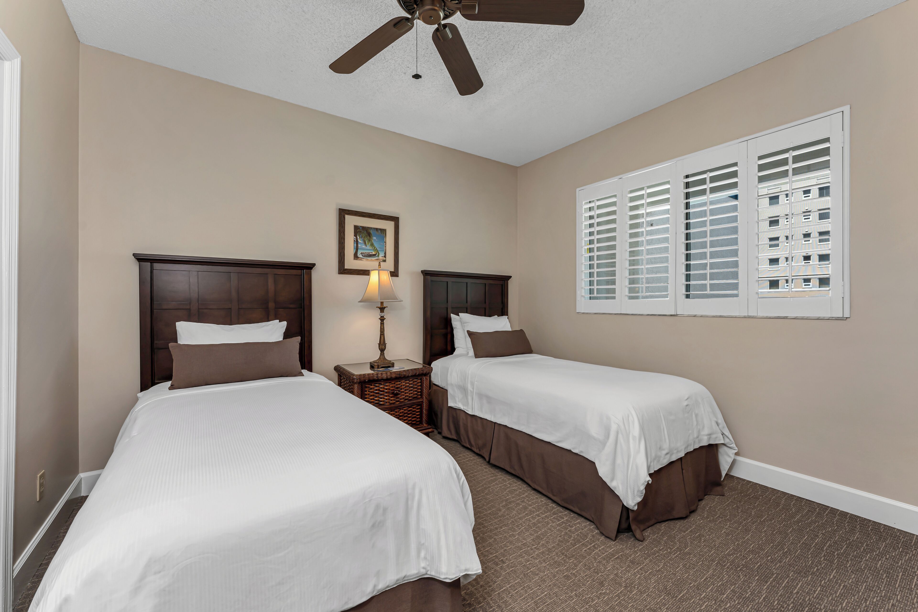 A hotel room with two beds, a ceiling fan, a lamp, and a window with blinds.