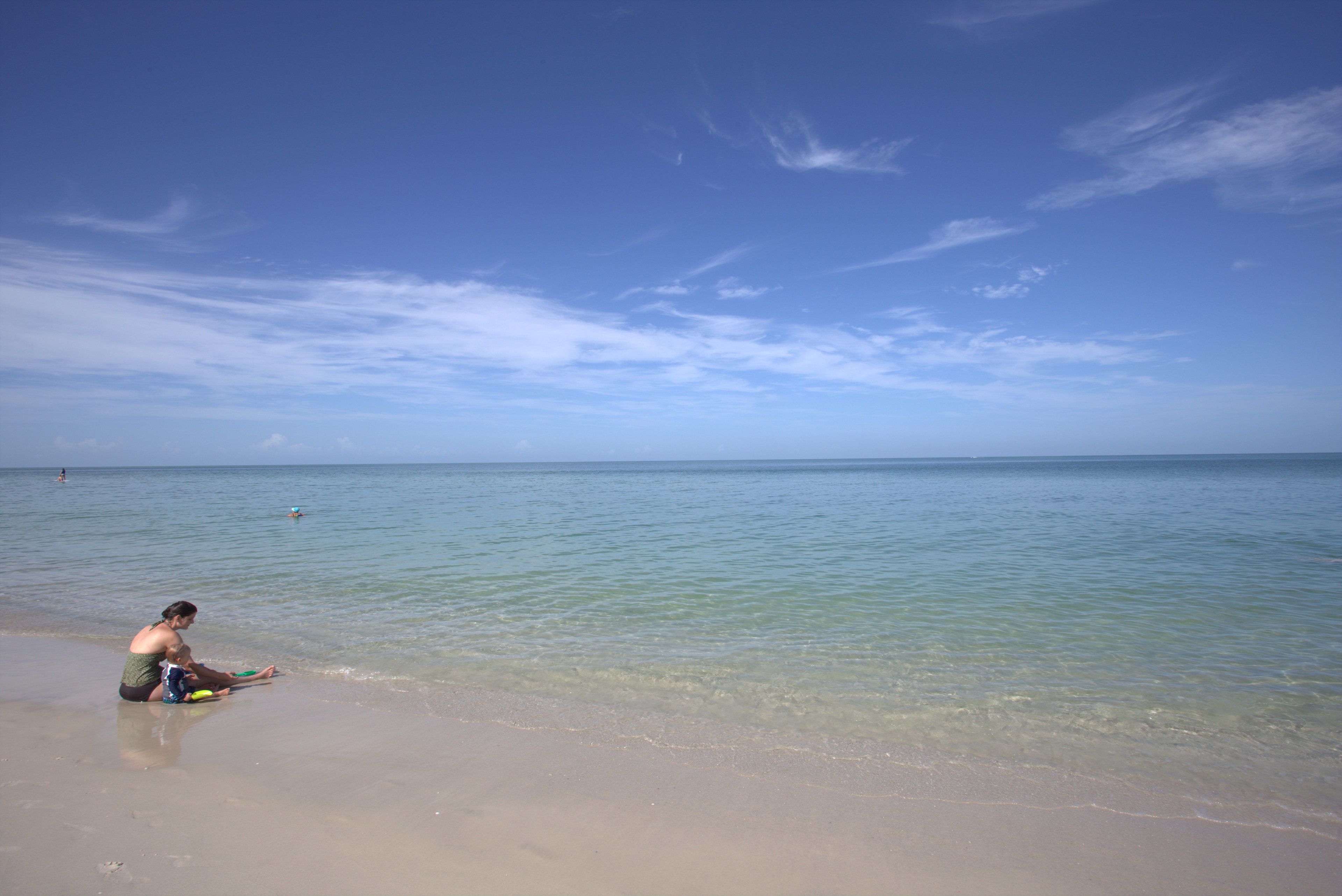 Een strand met helder water en iemand die op het zand zit.