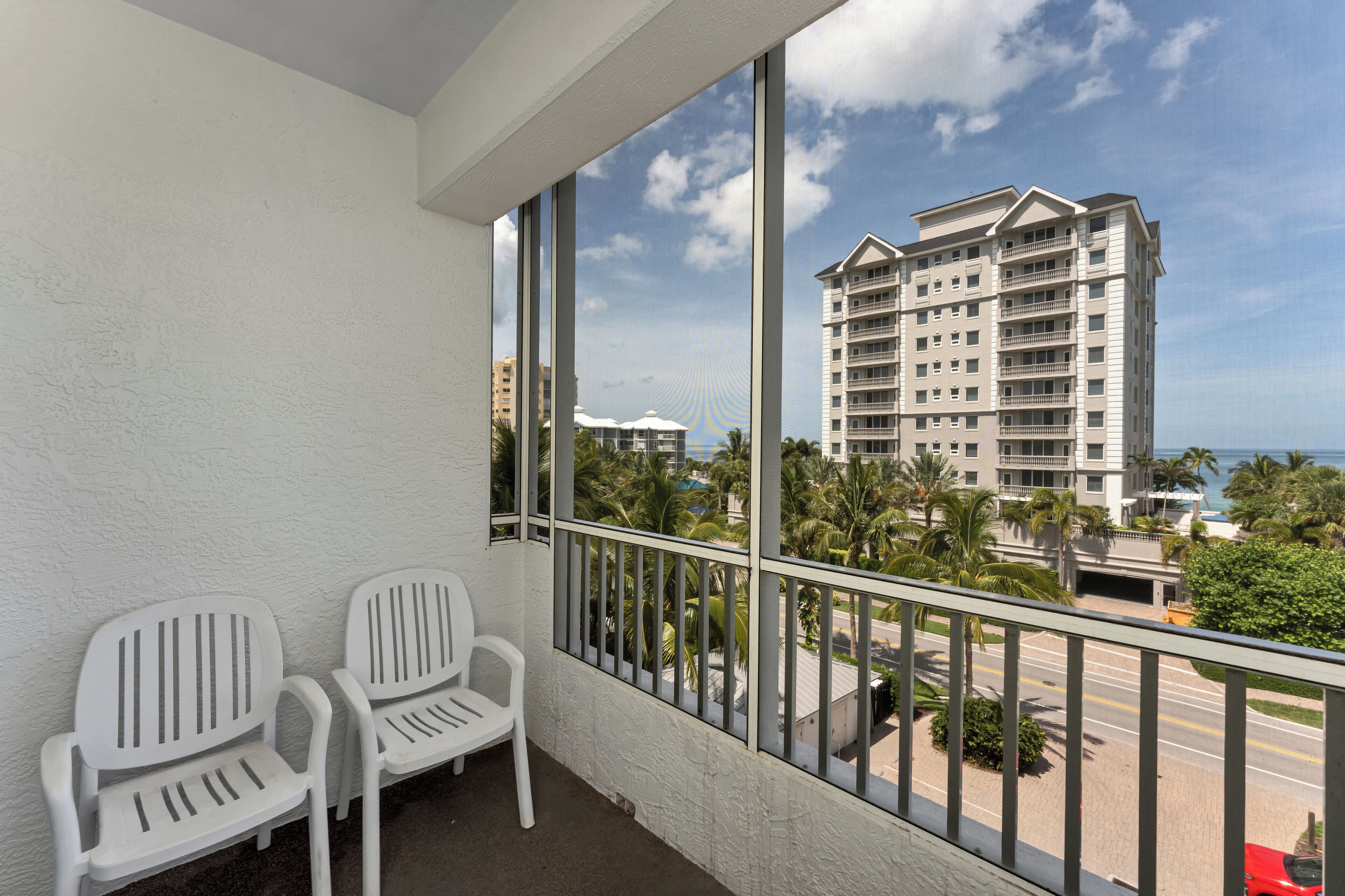 A balcony with two white chairs, a view of a tall building, palm trees, and a road.