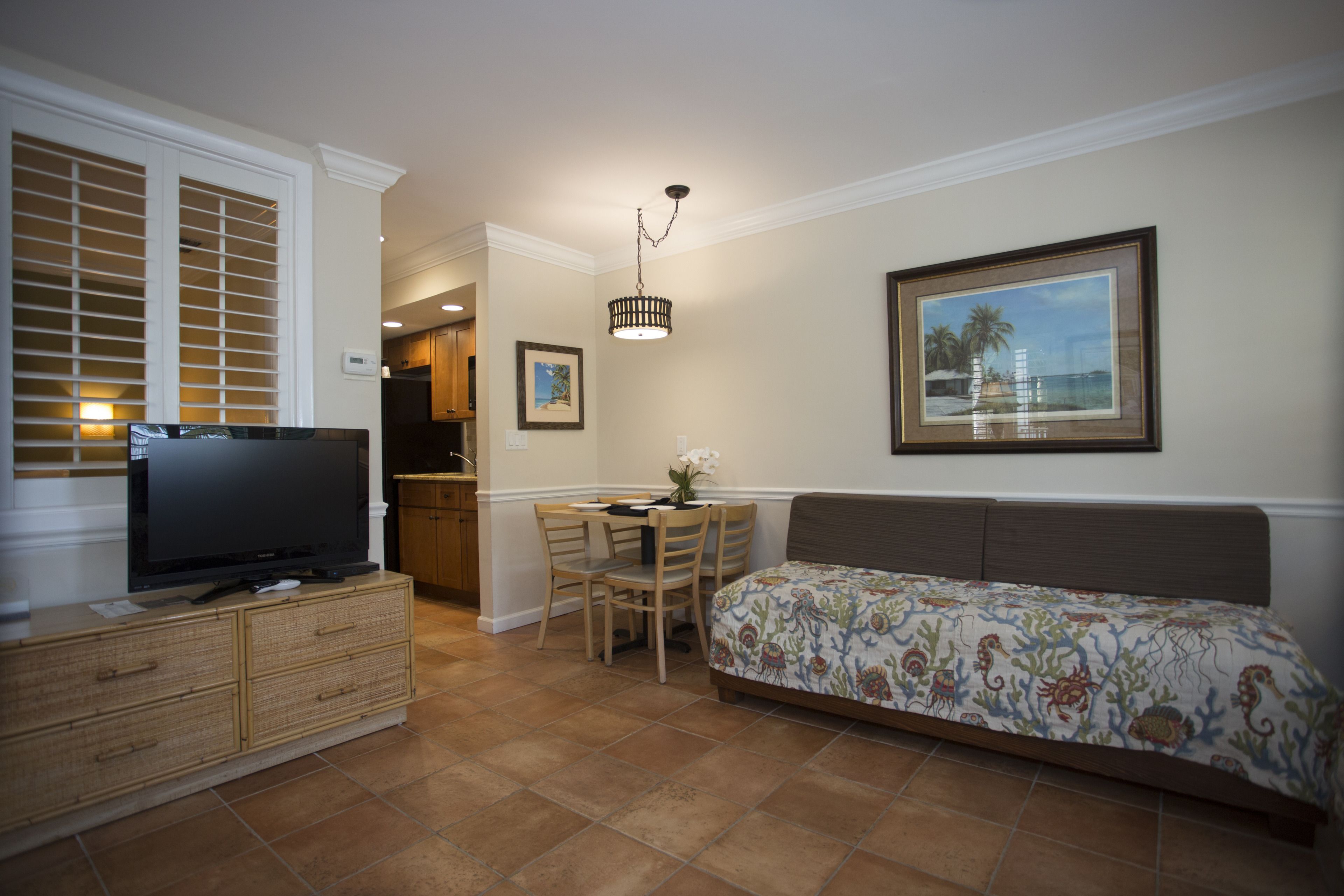 Une chambre avec un lit, un téléviseur posé sur une commode, une table à manger avec des chaises, et une cuisine à l’arrière-plan.