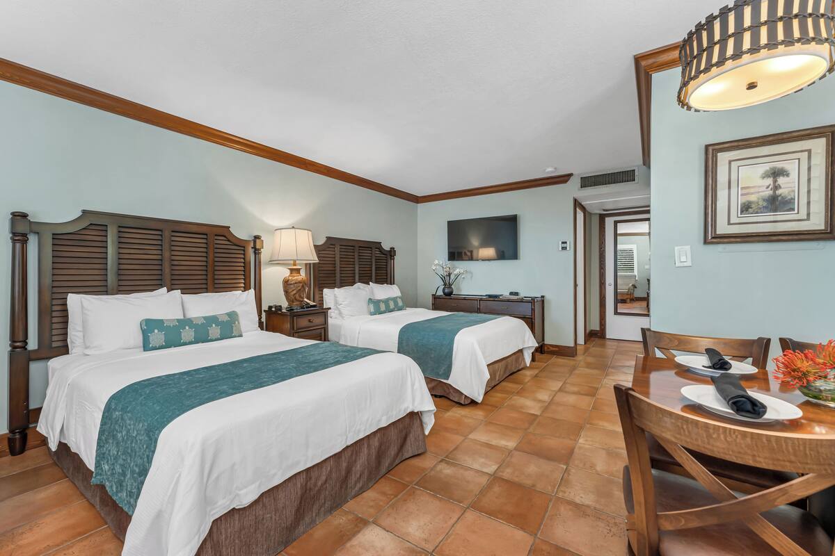 A hotel room with two beds, a dining table, chairs, a lamp, and a framed picture on the wall.