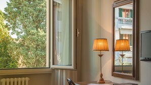 A room with a desk, two lamps, a radiator, and a window with a view of trees and a balcony. - Hotel La Pace (Pontedera)