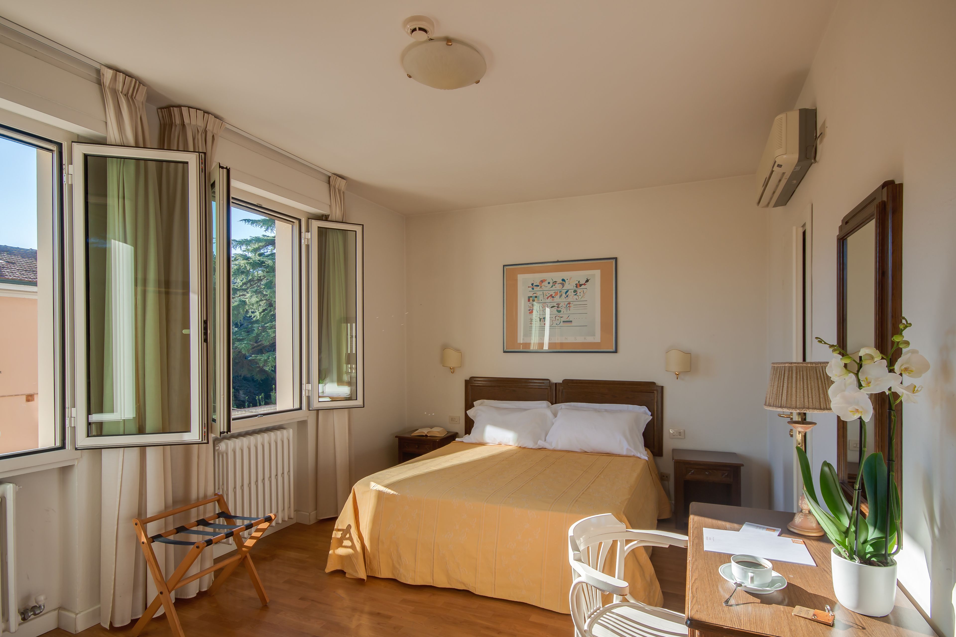 Ein Hotelzimmer mit Bett, Schreibtisch mit Lampe, Stuhl, Fenster mit Vorhängen, Heizkörper und Klimaanlage.