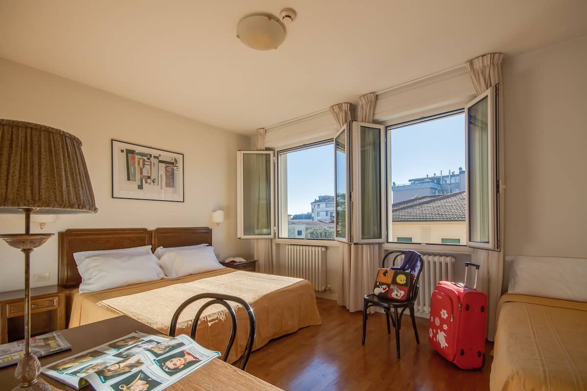 Une chambre d’hôtel avec un lit, une table avec des magazines, une chaise, une valise et une fenêtre donnant sur des immeubles.