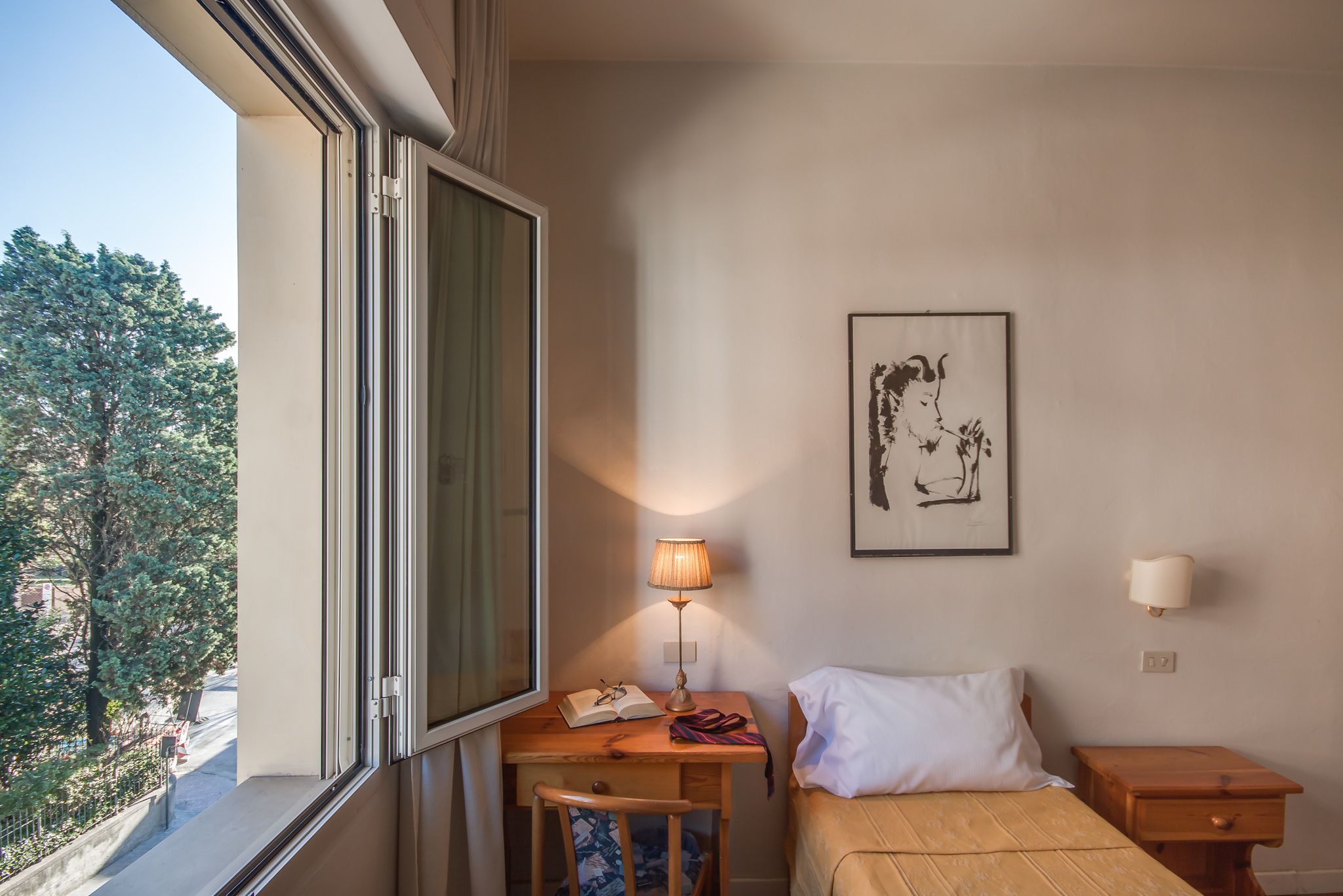 Ein Hotelzimmer mit Bett, Schreibtisch mit Lampe, Stuhl, Nachttisch und einem gerahmten Kunstwerk an der Wand.