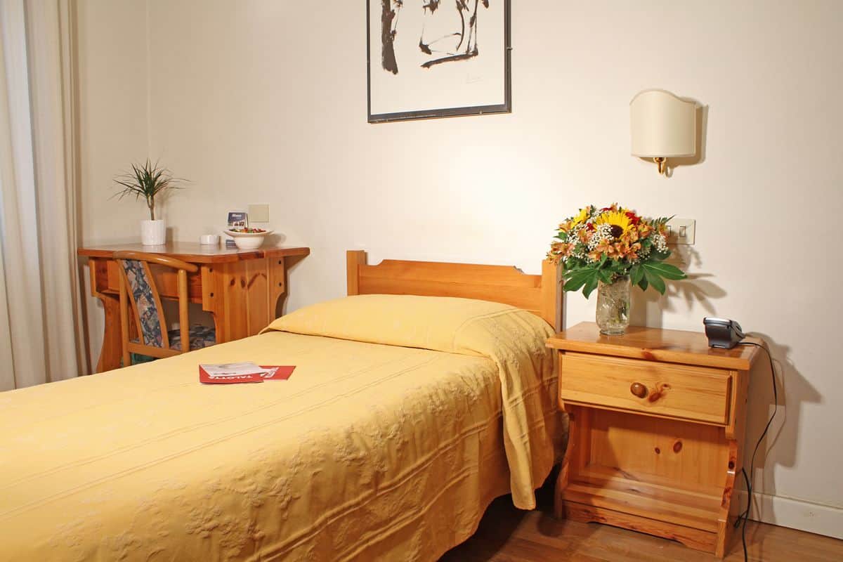 Une chambre bien rangée, avec un lit, une table de chevet, une lampe et un petit bureau agrémenté d’une plante.
